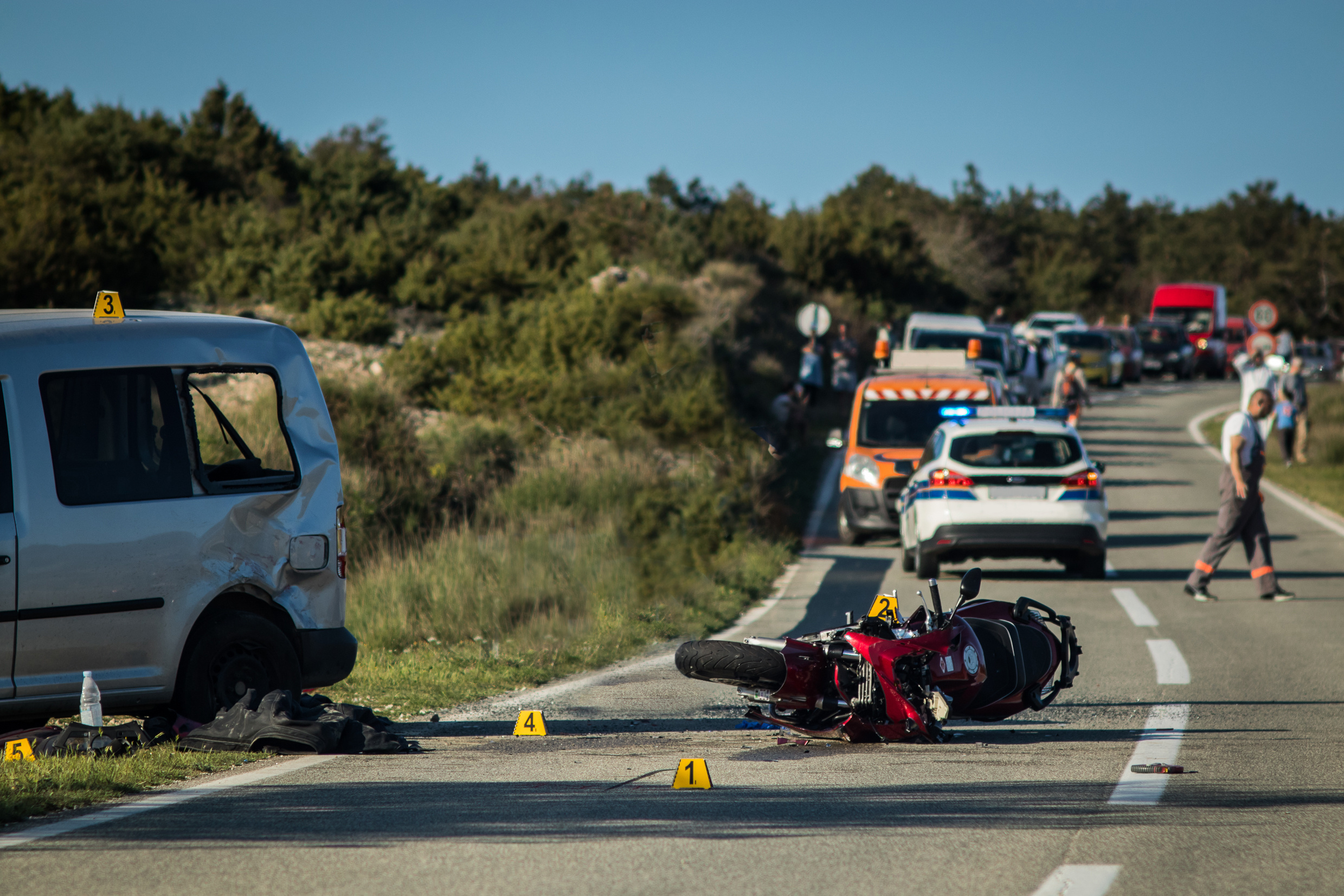 Car and a red sport motorcycle crash scene on an open road in afternoon. Workers and police seen around the crash site, with a queue of traffic building behind. Destroyed van and motorcycle.