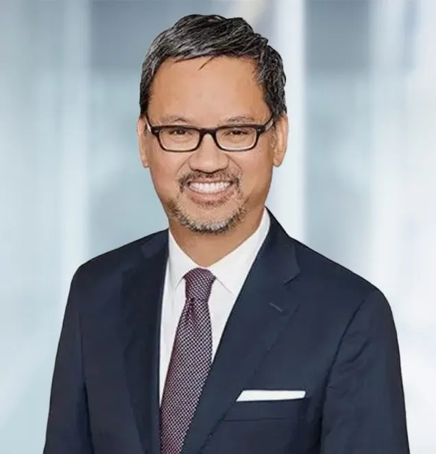 Professional headshot of a man in a dark suit, purple tie, and glasses smiling.