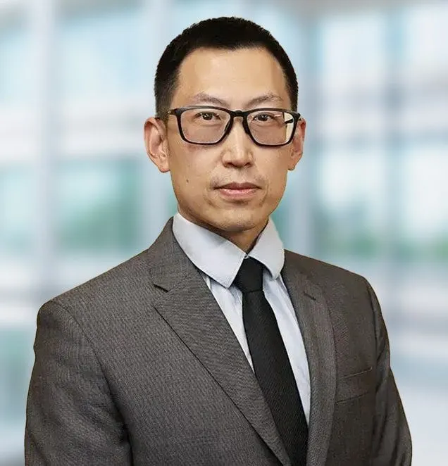 Professional headshot of a man in gray suit and black tie wearing glasses in office setting.