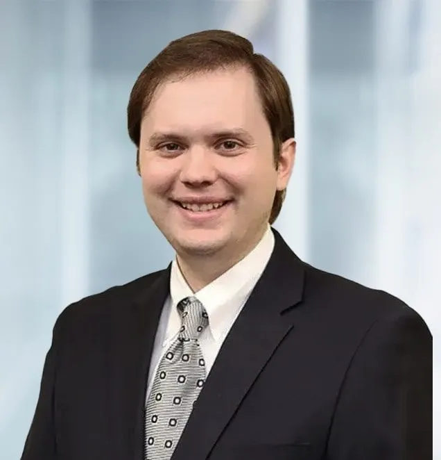 Professional headshot of a man in navy suit and patterned tie smiling in office setting.
