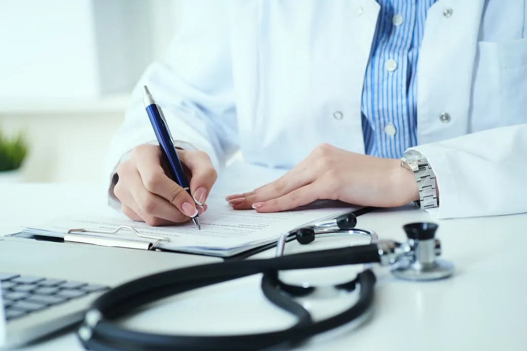 Mid section of female doctor writing prescription to patient at worktable.