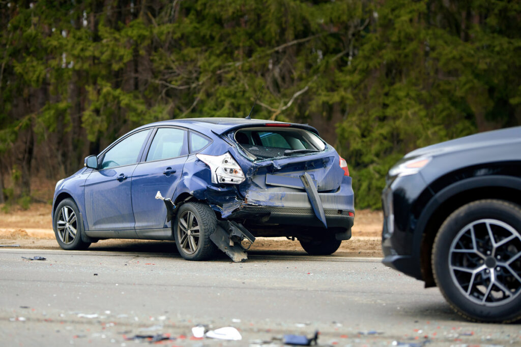 Blue compact car with severe rear end damage after highway collision, debris scattered on the road and another vehicle visible at the scene of the traffic accident. Damaged car with heavy rear impact
