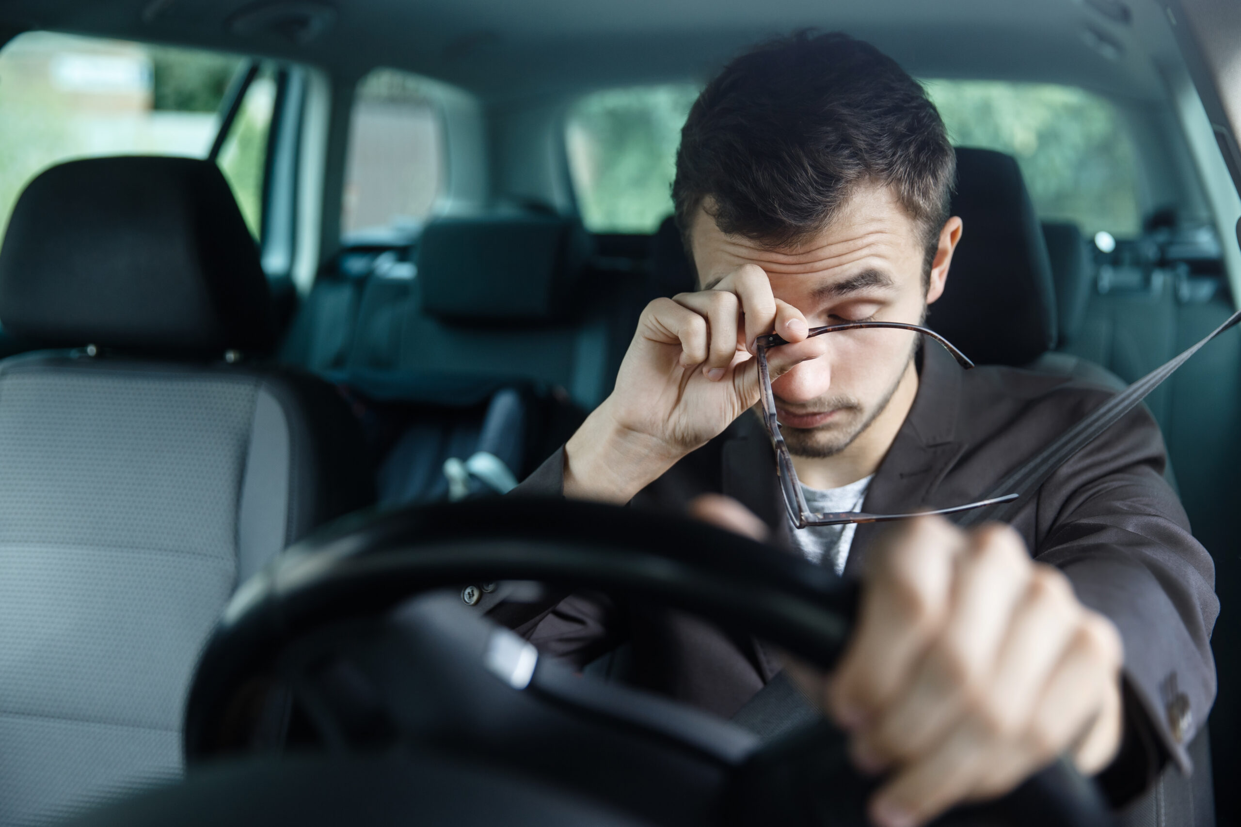 Sleepy young man rubbing his eyes with one hand while the other rests on the steering wheel