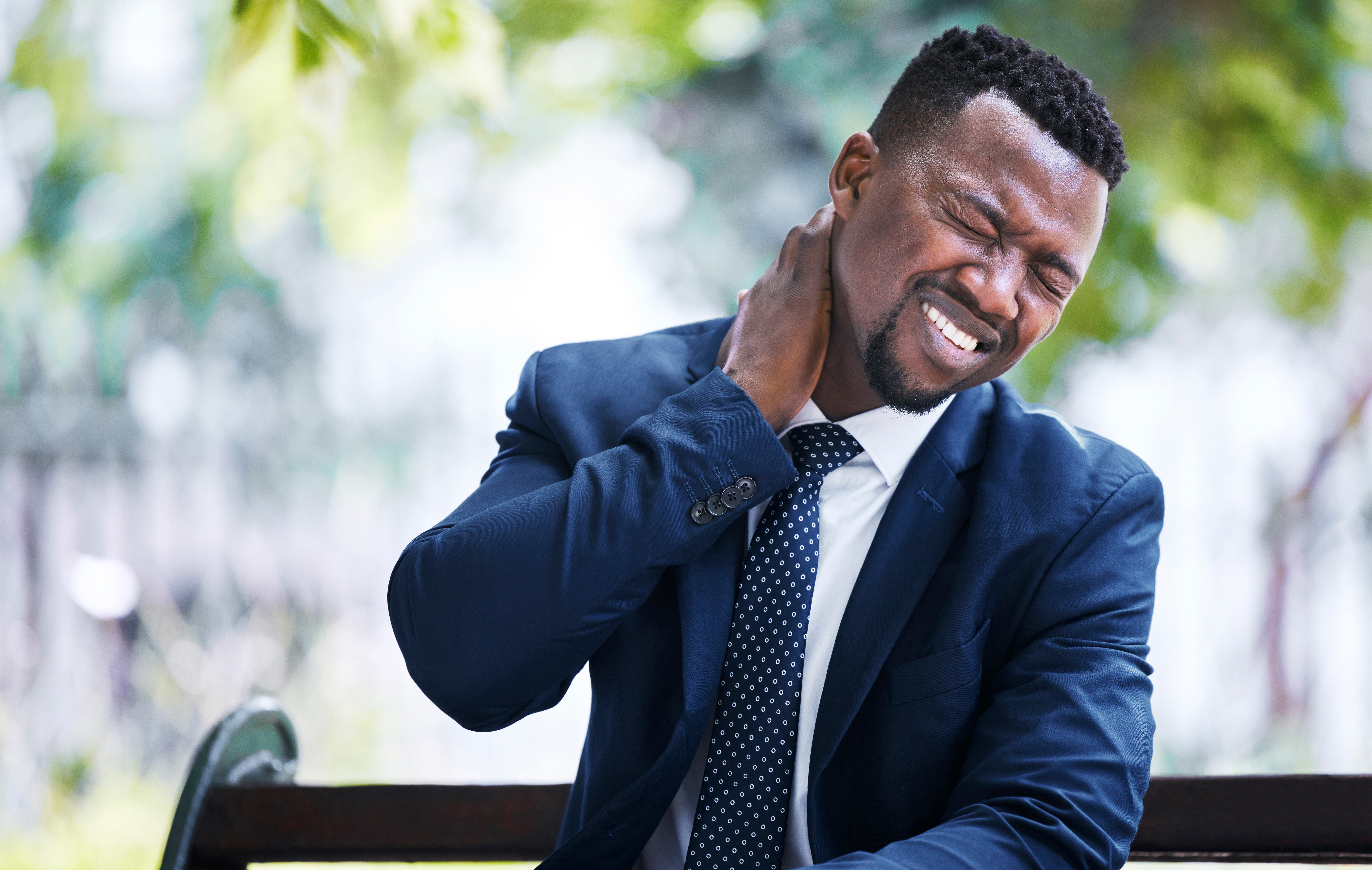 Business man holding his neck in pain while sitting on a bench after an accident