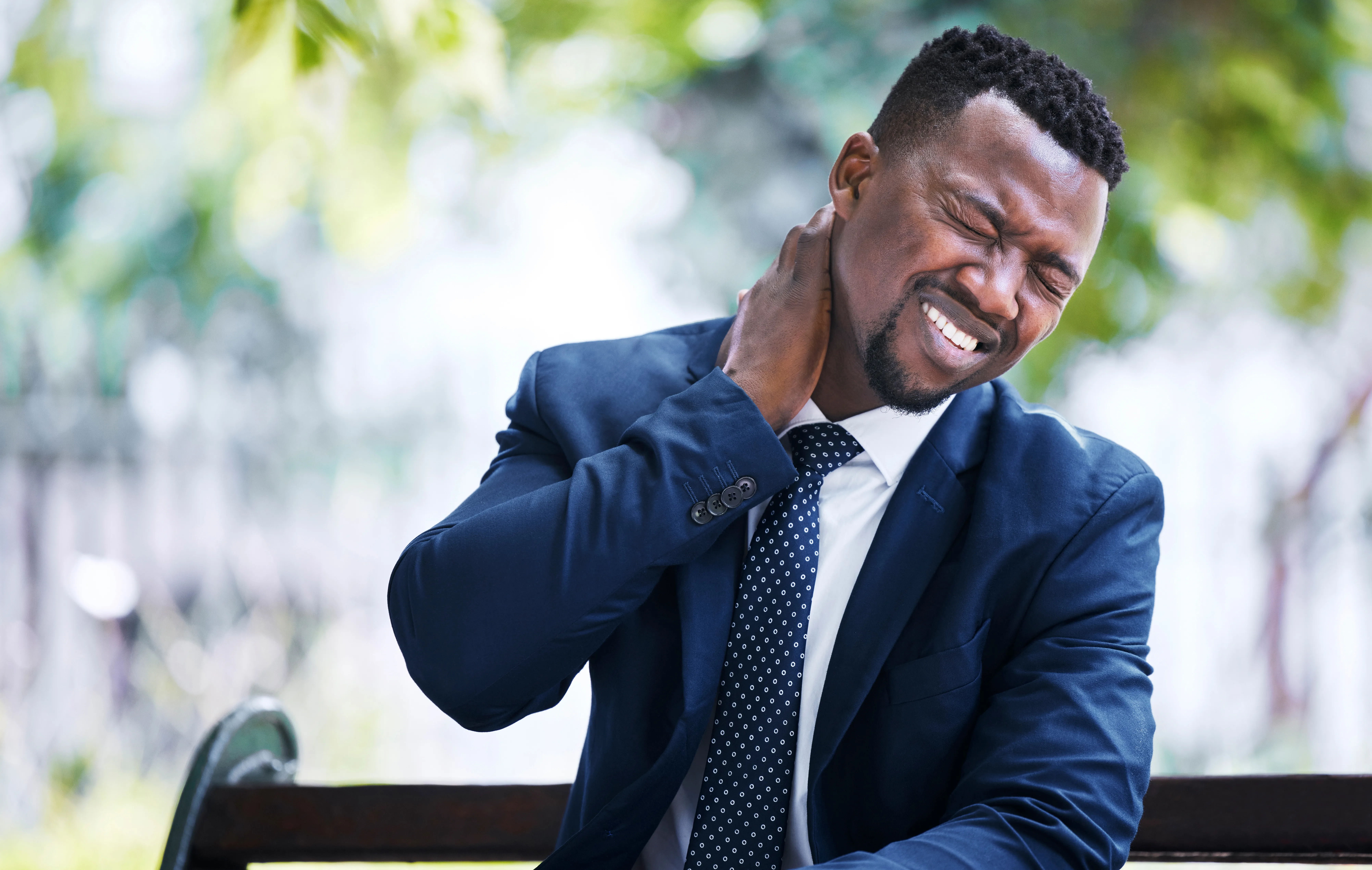 Business man holding his neck in pain while sitting on a bench after an accident