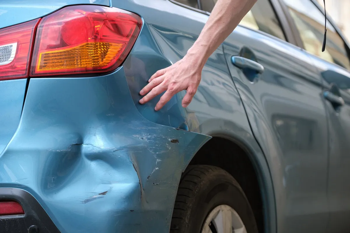 Driver hand examining dented car with damaged fender parked on city street side. Road safety and vehicle insurance concept